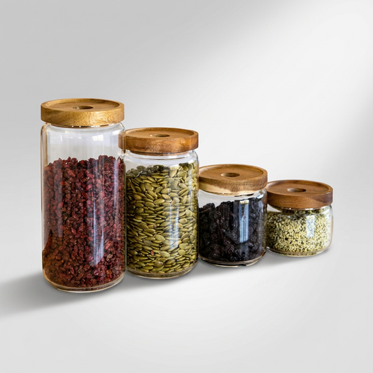 Four clear glass storage jars with light brown bamboo lids, arranged in descending size order. They are filled with various dried foods: red berries, green pumpkin seeds, dark raisins, and green tea leaves.
