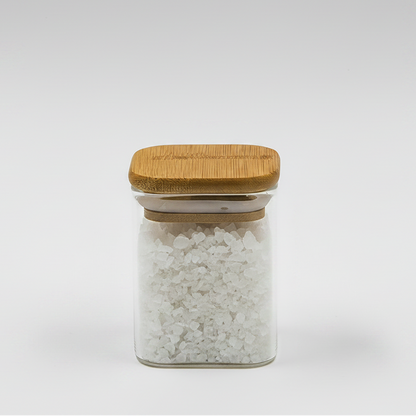 A clear glass storage jar with a bamboo lid, filled with white coarse salt or sugar crystals, shown on a light gray background with overlaid dimension lines indicating 60mm width and 80mm height. A walnut is placed to the left as a prop.