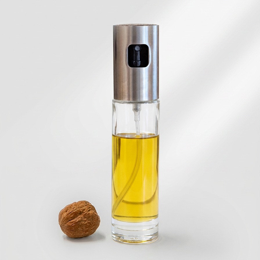 A clear glass oil spray bottle filled with golden yellow oil, topped with a brushed silver metal cap, shown on a light gray background next to a walnut.