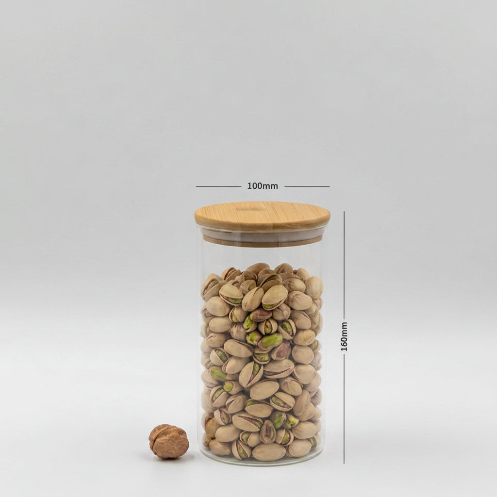 A clear glass jar with a light wood lid, filled with pistachios, shown on a light gray background with dimension lines indicating 100mm in diameter and 160mm in height. A walnut is placed beside the jar.
