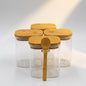 Four clear glass storage jars with square bamboo lids are arranged on a white surface, with one jar in front holding a matching bamboo spoon. A soft diagonal light beam casts shadows on the background.